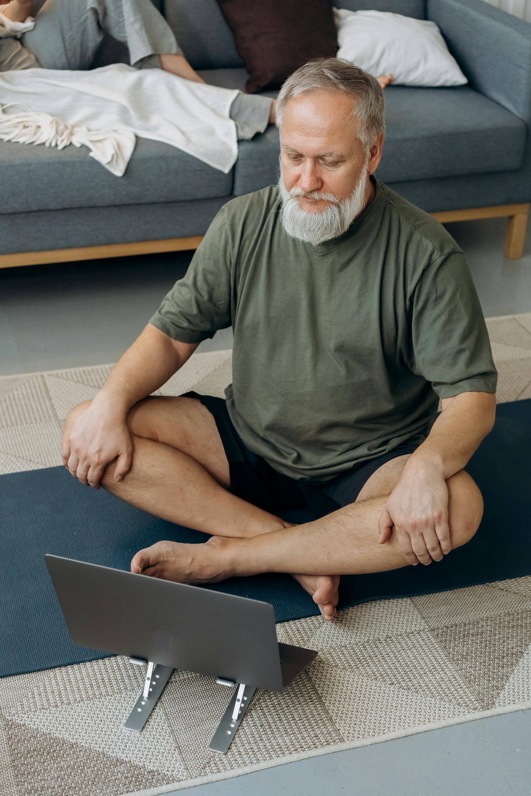 Pilates 2 Older man practicing yoga while following an online session on a laptop in his living room.
