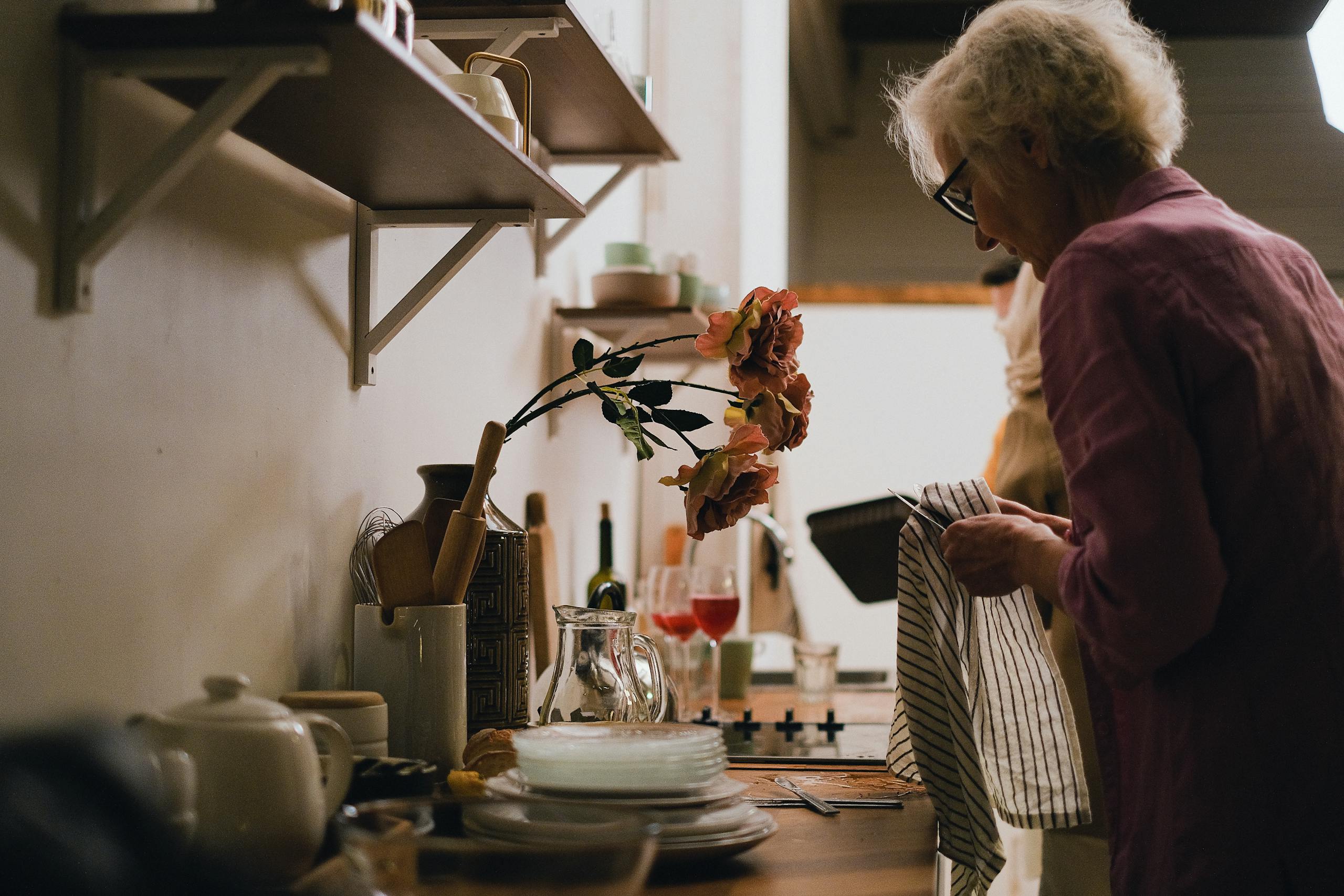 Elderly woman engaged in dish drying in a cozy, homey kitchen setting with shelves and flowers.